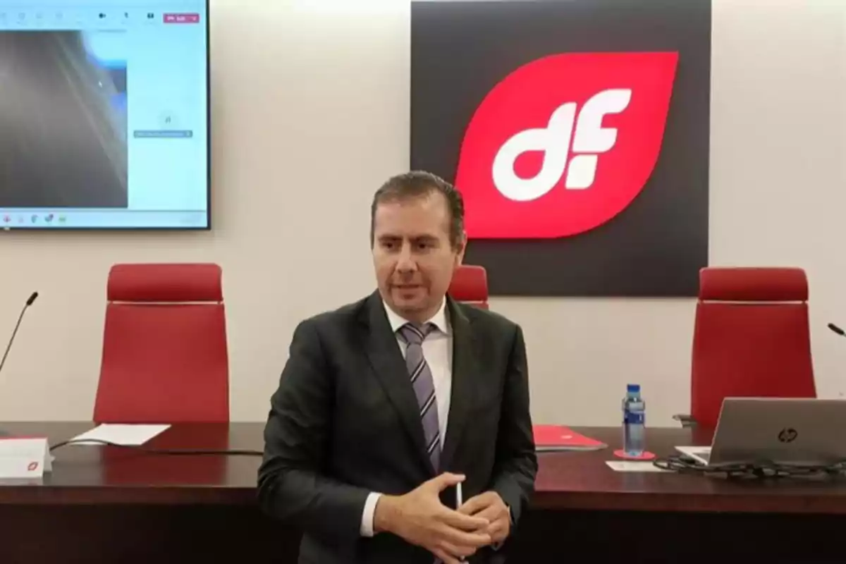 Hombre de traje en una sala de conferencias con sillas rojas, una pantalla y un logotipo rojo con las letras df al fondo Hombre de traje en una sala de conferencias con sillas rojas, una pantalla y un logotipo rojo con las letras df al fondo