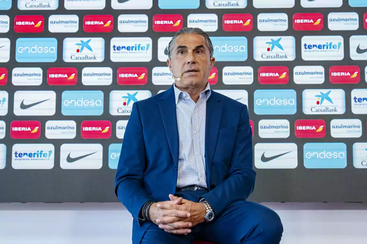 Hombre de traje azul sentado frente a un fondo con logotipos de patrocinadores deportivos