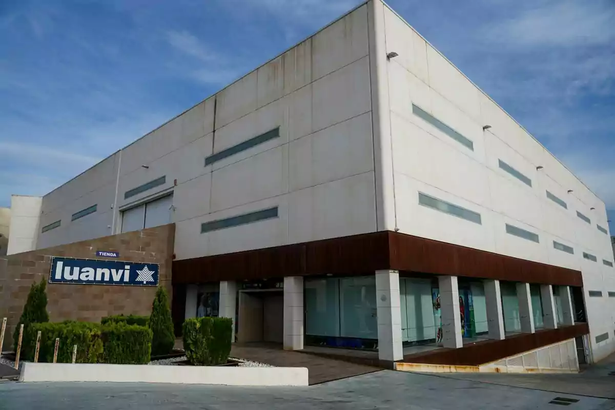 Edificio moderno de color blanco con ventanas alargadas y un letrero que dice luanvi en la entrada rodeado de arbustos y cielo despejado