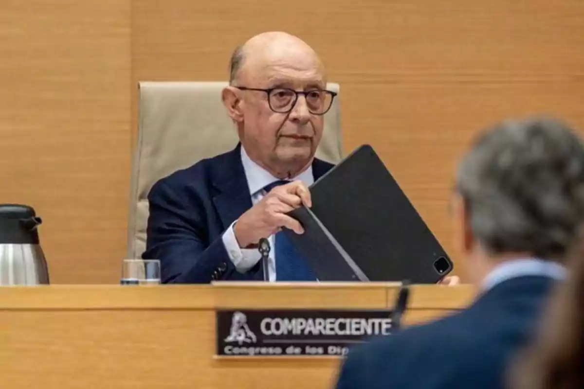 Hombre calvo con gafas y traje sosteniendo una tablet en una sala de reuniones oficiales con un cartel que dice compareciente Hombre calvo con gafas y traje sosteniendo una tablet en una sala de reuniones oficiales con un cartel que dice compareciente