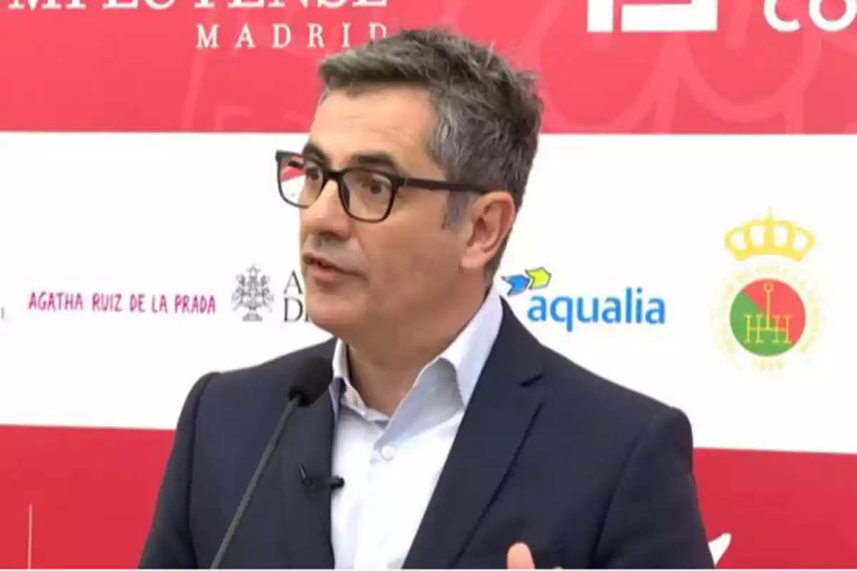 Hombre de cabello canoso y gafas hablando frente a un micrófono en un evento con fondo de logotipos y texto. Hombre de cabello canoso y gafas hablando frente a un micrófono en un evento con fondo de logotipos y texto.