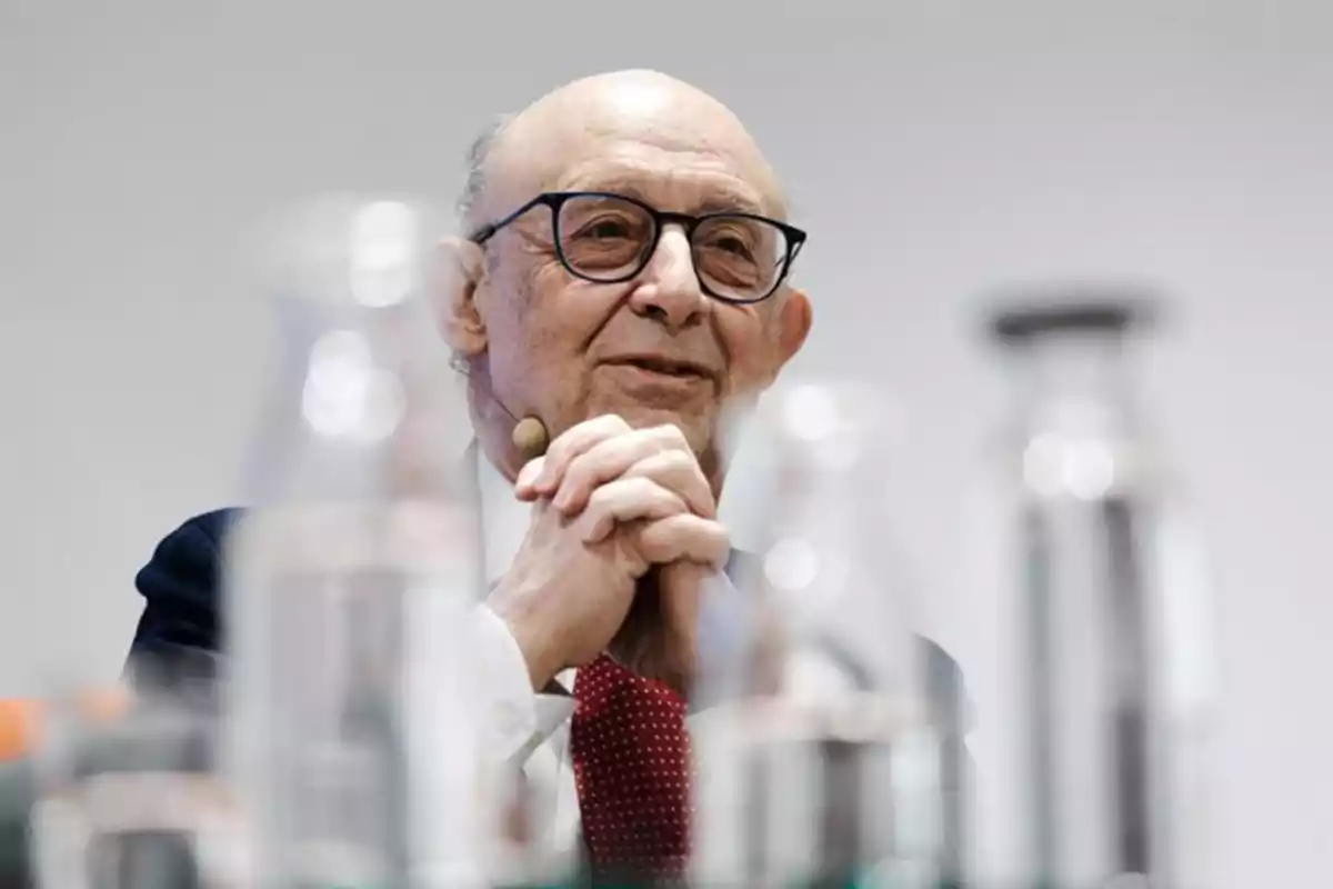Hombre mayor con gafas y corbata roja hablando en una conferencia con botellas de vidrio desenfocadas en primer plano