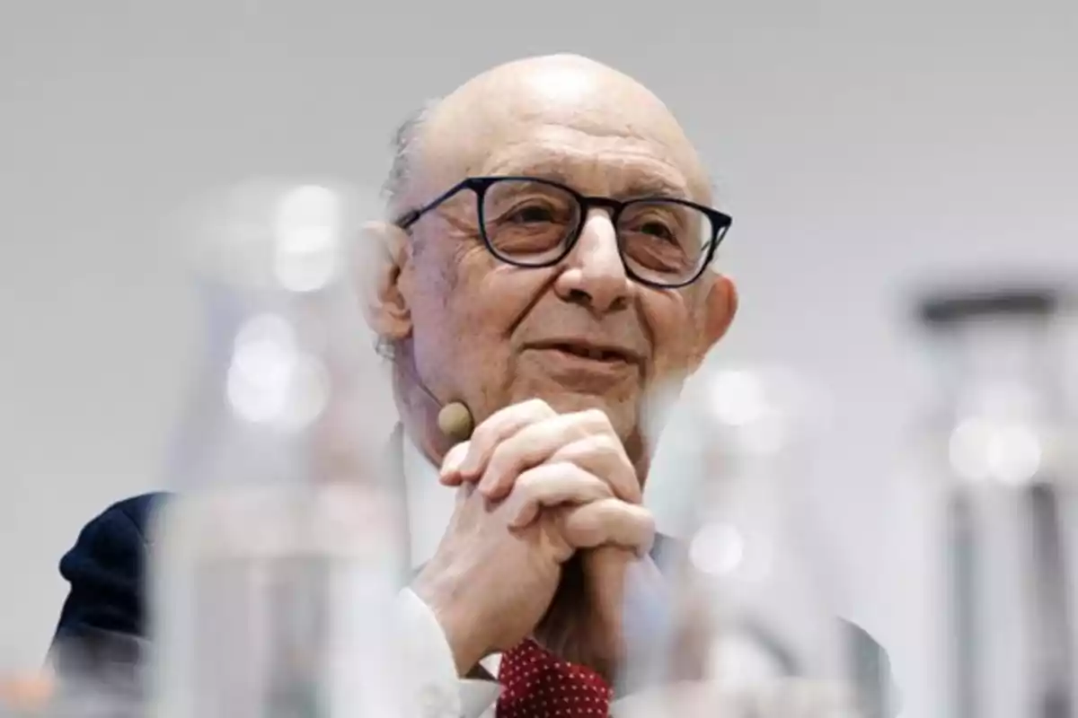 Hombre mayor con gafas y micrófono de diadema hablando en un evento, con las manos entrelazadas y botellas desenfocadas en primer plano Hombre mayor con gafas y micrófono de diadema hablando en un evento, con las manos entrelazadas y botellas desenfocadas en primer plano