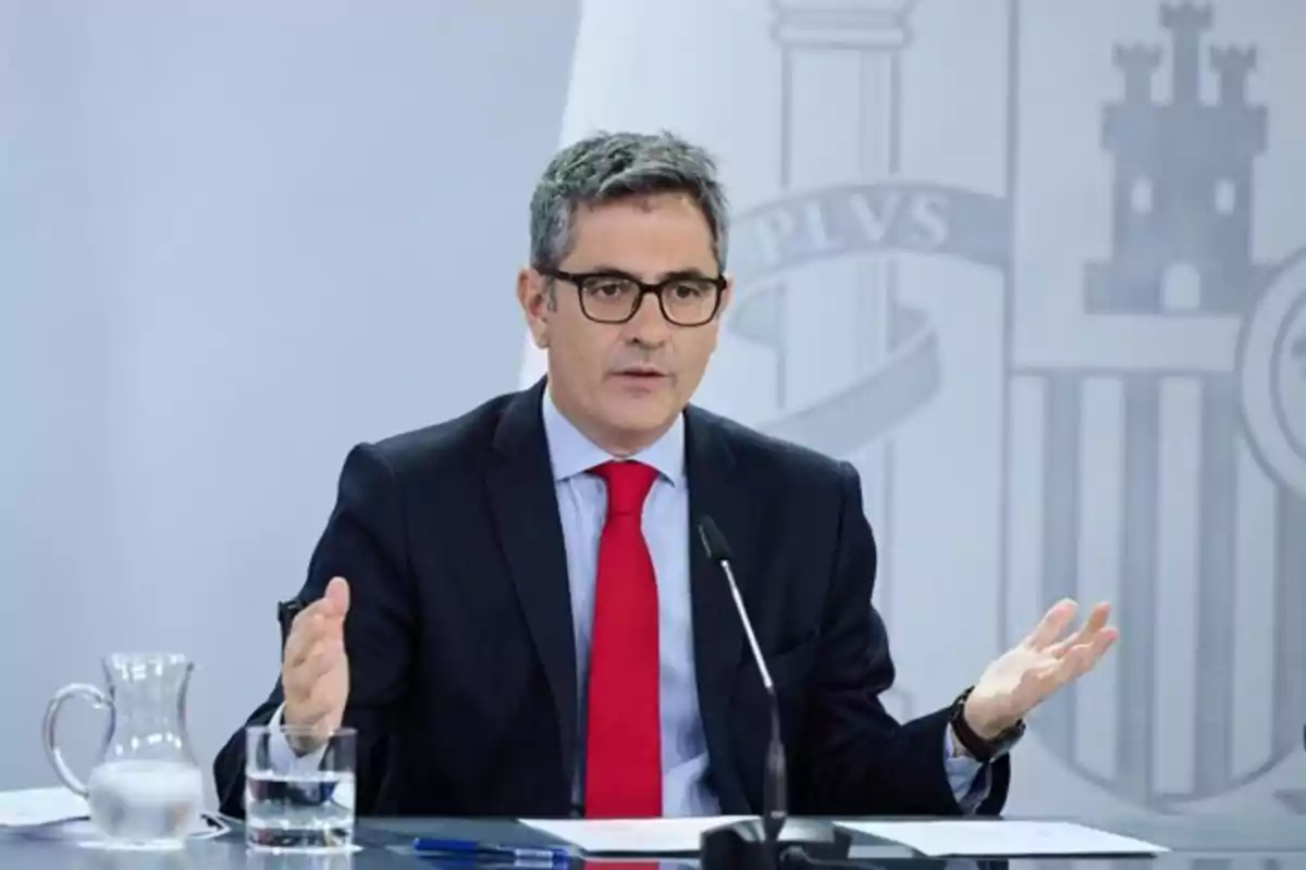 Hombre de cabello canoso y gafas con traje oscuro y corbata roja hablando en una conferencia de prensa sentado frente a un micrófono con una jarra y un vaso de agua sobre la mesa y un fondo institucional Hombre de cabello canoso y gafas con traje oscuro y corbata roja hablando en una conferencia de prensa sentado frente a un micrófono con una jarra y un vaso de agua sobre la mesa y un fondo institucional