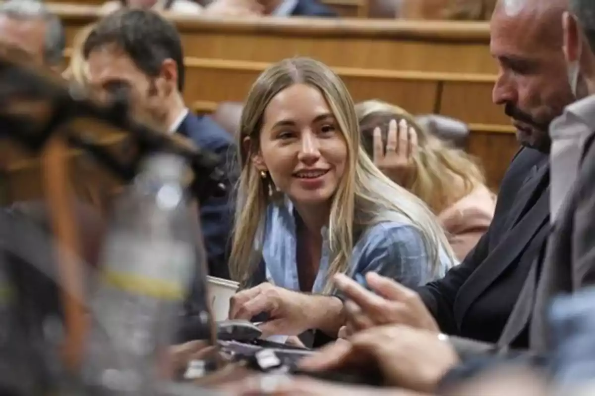 Una mujer joven de cabello rubio y largo sonríe mientras conversa con un hombre en un recinto parlamentario rodeada de otras personas Una mujer joven de cabello rubio y largo sonríe mientras conversa con un hombre en un recinto parlamentario rodeada de otras personas