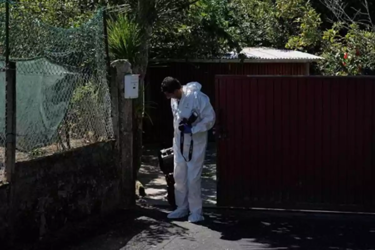 Persona con traje de protección blanca y guantes azules sostiene equipo especializado mientras ingresa a una propiedad cercada con portón rojo