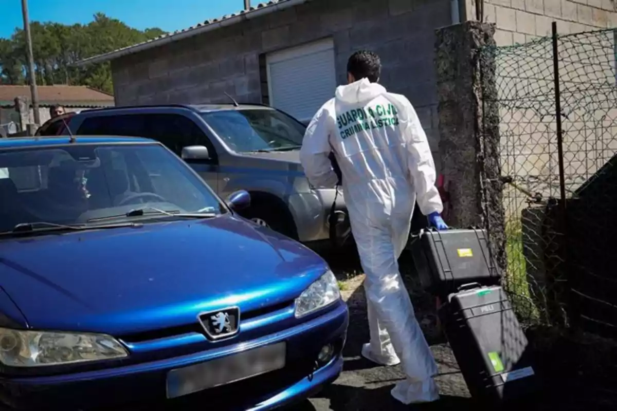 Un agente forense de la Guardia Civil con traje blanco y guantes azules transporta dos maletines negros junto a un coche azul estacionado frente a una vivienda Un agente forense de la Guardia Civil con traje blanco y guantes azules transporta dos maletines negros junto a un coche azul estacionado frente a una vivienda