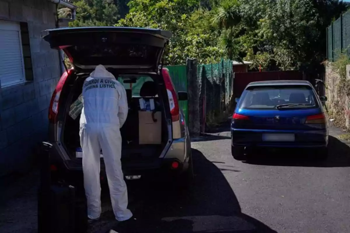 Persona con mono blanco de la Guardia Civil inspeccionando el maletero de un coche gris estacionado junto a una casa, con otro coche azul aparcado cerca y vegetación al fondo Persona con mono blanco de la Guardia Civil inspeccionando el maletero de un coche gris estacionado junto a una casa, con otro coche azul aparcado cerca y vegetación al fondo