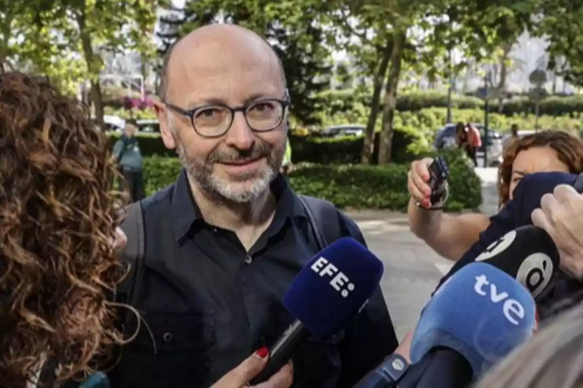 Un hombre con gafas y barba es entrevistado por varios periodistas al aire libre rodeado de micrófonos de diferentes medios de comunicación Un hombre con gafas y barba es entrevistado por varios periodistas al aire libre rodeado de micrófonos de diferentes medios de comunicación