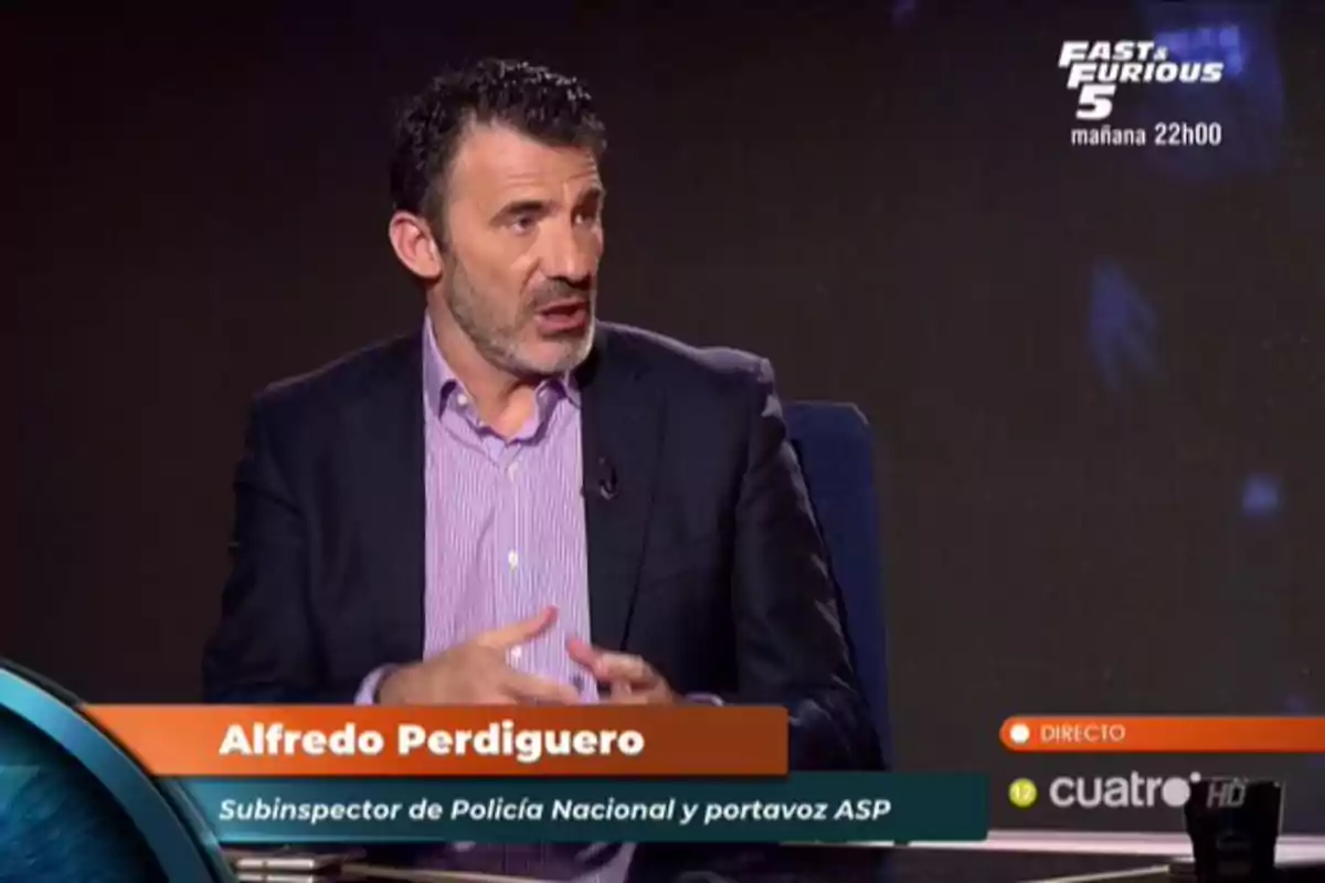 Hombre de cabello corto y barba, vestido con saco oscuro y camisa lila, hablando en un programa de televisión con rótulo que indica su nombre y cargo, en un set con fondo oscuro y gráficos de canal en pantalla.