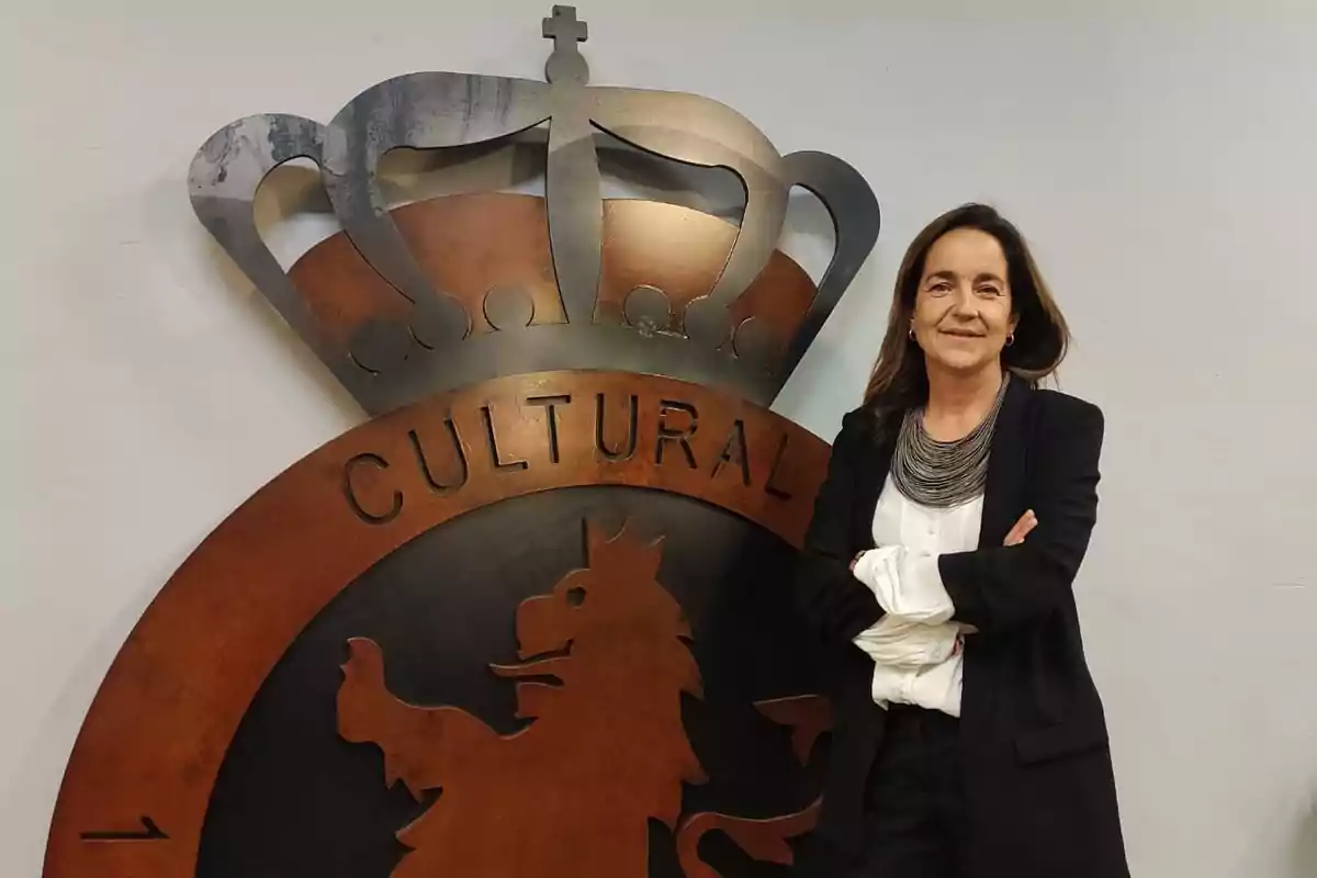 Mujer de cabello castaño y vestimenta formal posando sonriente con los brazos cruzados frente a un gran escudo metálico con la palabra CULTURAL y una corona Mujer de cabello castaño y vestimenta formal posando sonriente con los brazos cruzados frente a un gran escudo metálico con la palabra CULTURAL y una corona