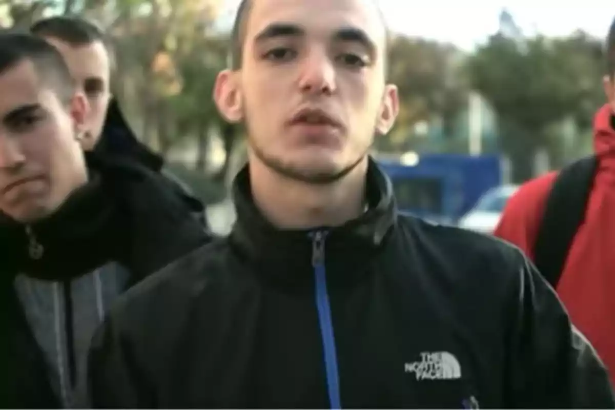 Joven con chaqueta negra de The North Face mirando a la cámara mientras otras personas aparecen desenfocadas al fondo en un entorno urbano Joven con chaqueta negra de The North Face mirando a la cámara mientras otras personas aparecen desenfocadas al fondo en un entorno urbano