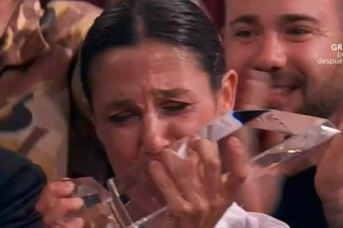 Gabriela emocionada sostiene un trofeo de cristal de MasterChef mientras llora de felicidad rodeada de personas que la apoyan.