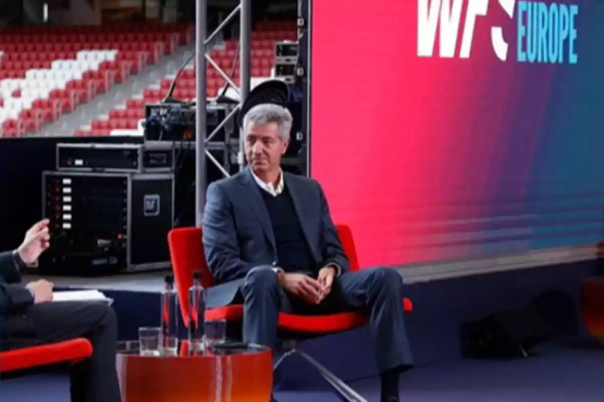 Hombre de cabello canoso con traje sentado en una silla roja durante un evento en un escenario con fondo de pantalla grande y letras WFS Europe Hombre de cabello canoso con traje sentado en una silla roja durante un evento en un escenario con fondo de pantalla grande y letras WFS Europe
