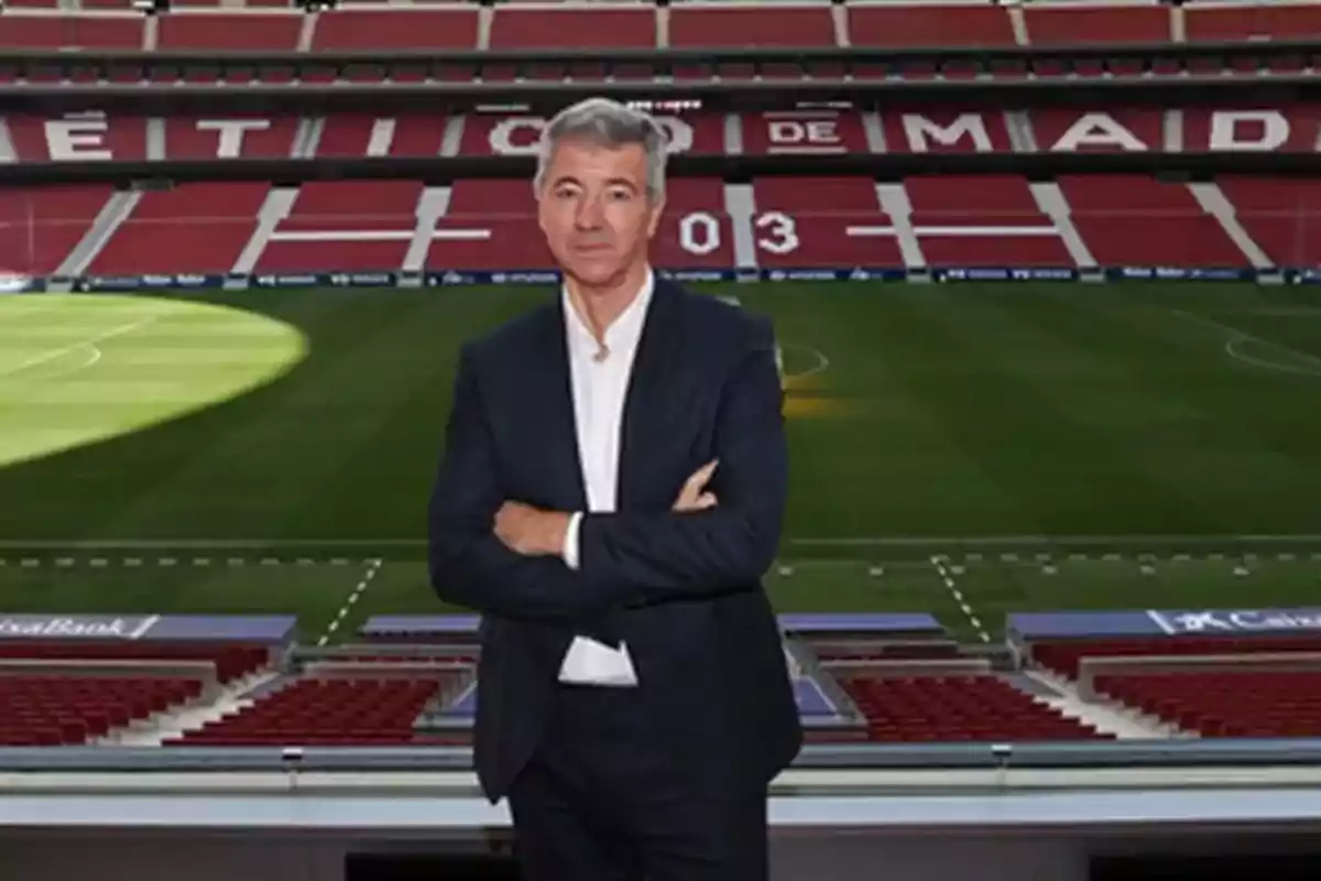 Hombre de cabello canoso con traje oscuro y camisa blanca posando con los brazos cruzados en un estadio de fútbol vacío
