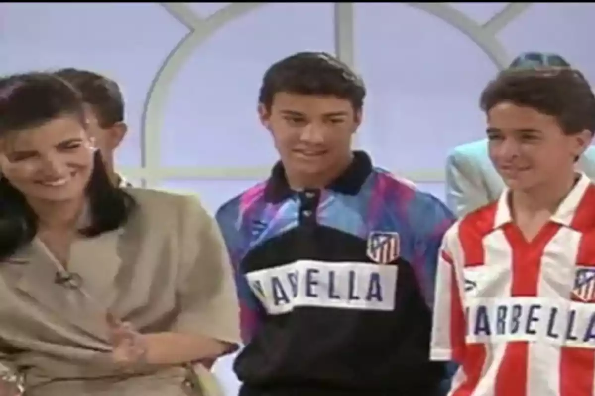 Tres personas sonríen en un set de televisión, dos de ellas llevan camisetas del Atlético de Madrid de los años noventa Tres personas sonríen en un set de televisión, dos de ellas llevan camisetas del Atlético de Madrid de los años noventa