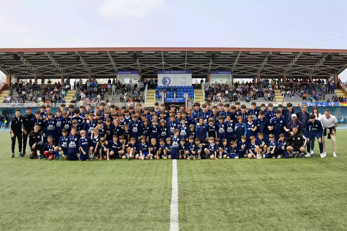 Un gran grupo de niños y jóvenes con uniforme azul posan para una foto en un campo de fútbol con gradas llenas de espectadores al fondo Un gran grupo de niños y jóvenes con uniforme azul posan para una foto en un campo de fútbol con gradas llenas de espectadores al fondo