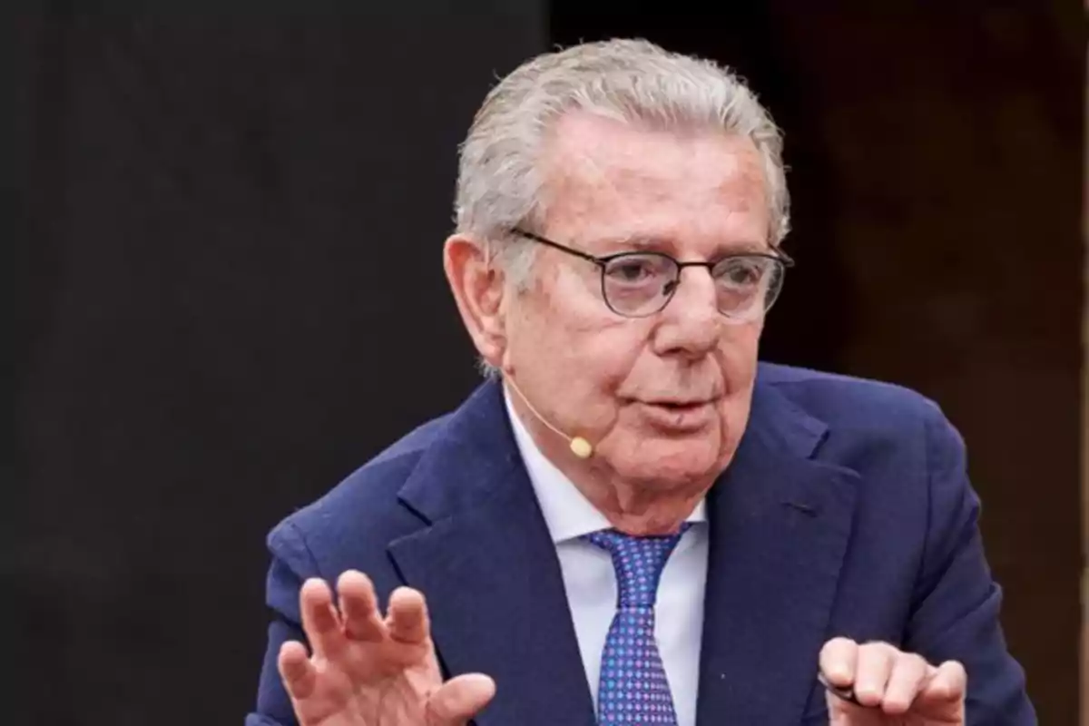 Hombre mayor con gafas y micrófono de diadema hablando y gesticulando con las manos mientras viste traje azul y corbata Hombre mayor con gafas y micrófono de diadema hablando y gesticulando con las manos mientras viste traje azul y corbata