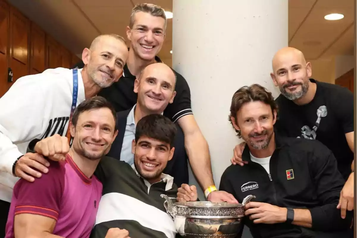 Grupo de seis hombres sonríen y posan juntos en un vestuario mientras uno de ellos sostiene un trofeo plateado grande