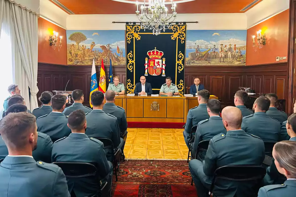 Un grupo de personas con uniformes militares asiste a una ceremonia oficial en una sala decorada con murales históricos, banderas y un escudo nacional, mientras varias autoridades están sentadas en una mesa al frente. Un grupo de personas con uniformes militares asiste a una ceremonia oficial en una sala decorada con murales históricos, banderas y un escudo nacional, mientras varias autoridades están sentadas en una mesa al frente.