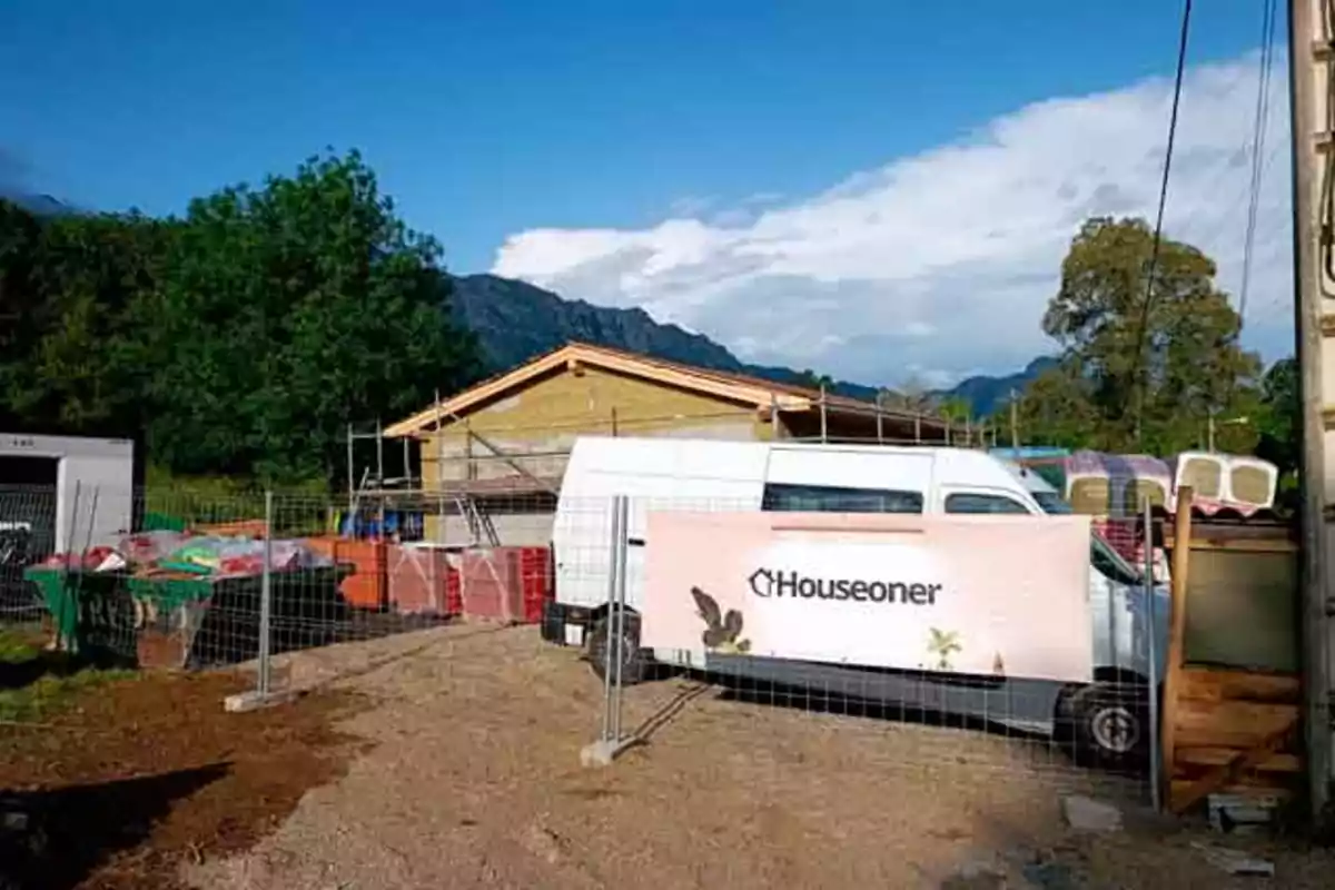 Obra en construcción con una furgoneta blanca estacionada frente a una casa de madera y un cartel de Houseoner, rodeada de materiales y árboles al fondo