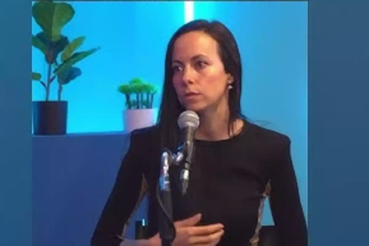 Mujer de cabello largo y oscuro hablando frente a un micrófono en un estudio con fondo azul y plantas decorativas Mujer de cabello largo y oscuro hablando frente a un micrófono en un estudio con fondo azul y plantas decorativas