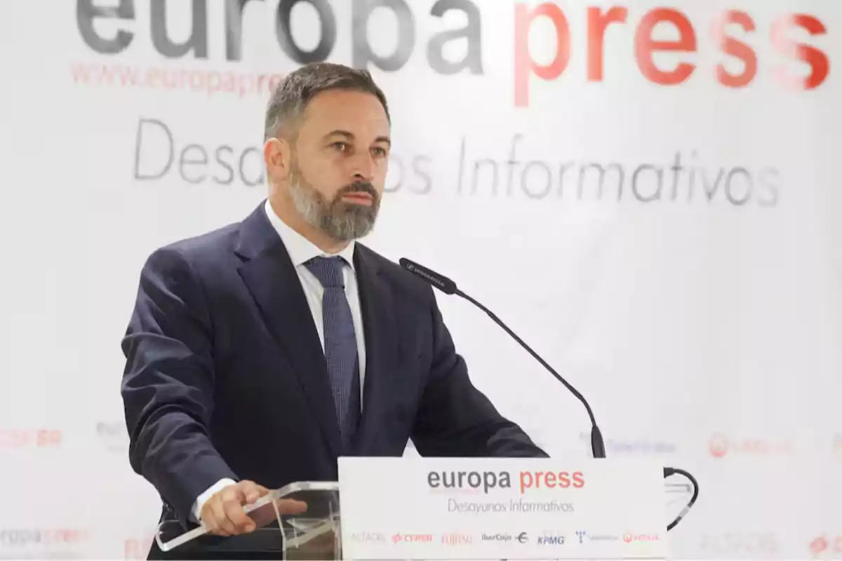 Hombre con barba y traje azul hablando en un atril durante un evento de Europa Press con fondo blanco y logotipos de patrocinadores. Hombre con barba y traje azul hablando en un atril durante un evento de Europa Press con fondo blanco y logotipos de patrocinadores.