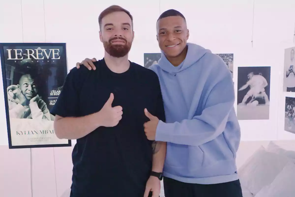 Dos personas posan sonrientes frente a una pared decorada con fotos y una revista, ambas hacen el gesto de pulgar arriba Dos personas posan sonrientes frente a una pared decorada con fotos y una revista, ambas hacen el gesto de pulgar arriba