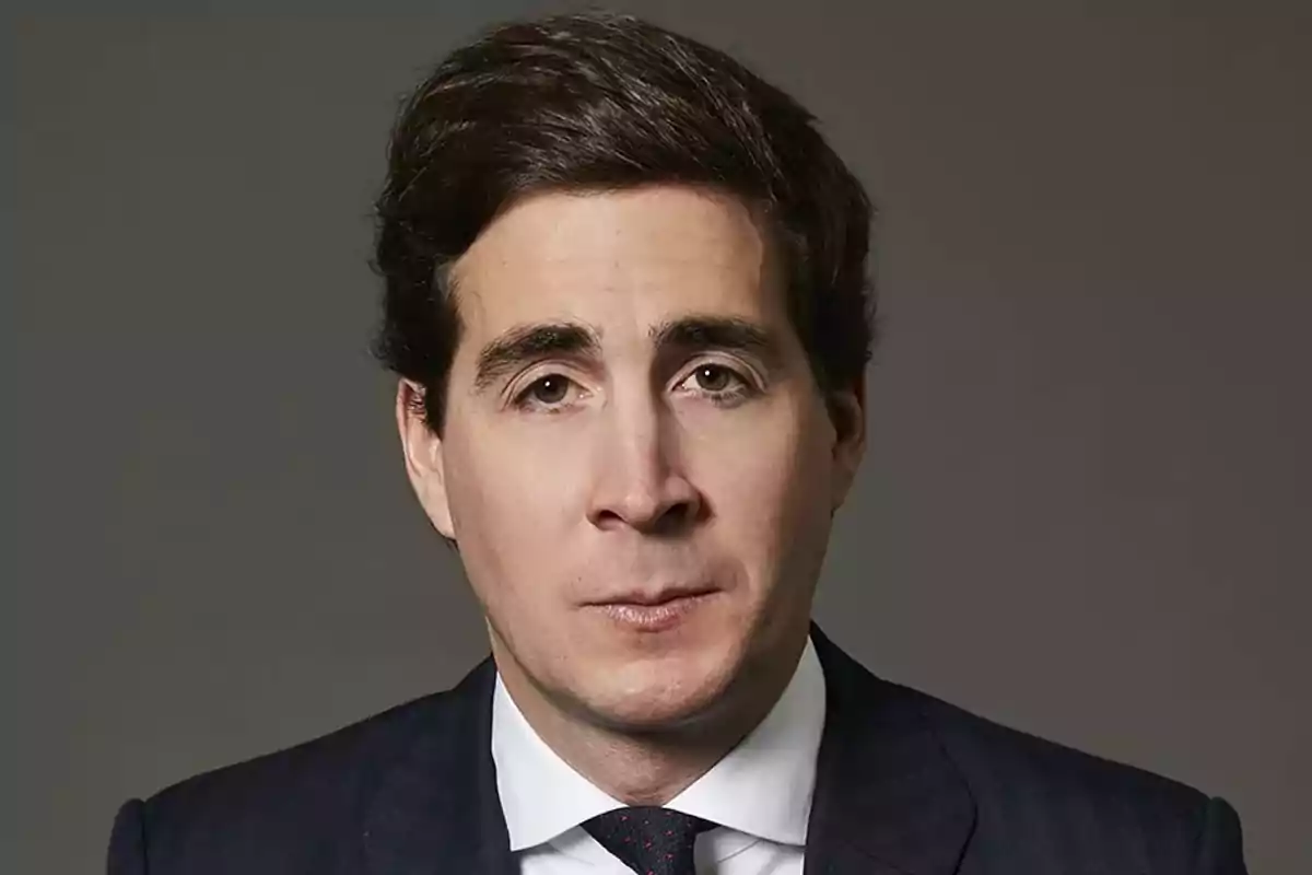 Hombre de cabello oscuro con traje azul marino y corbata, posando frente a un fondo gris Hombre de cabello oscuro con traje azul marino y corbata, posando frente a un fondo gris