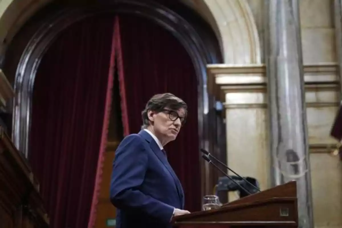 Hombre con gafas y traje azul hablando en un podio dentro de un edificio con cortinas rojas y columnas de mármol Hombre con gafas y traje azul hablando en un podio dentro de un edificio con cortinas rojas y columnas de mármol