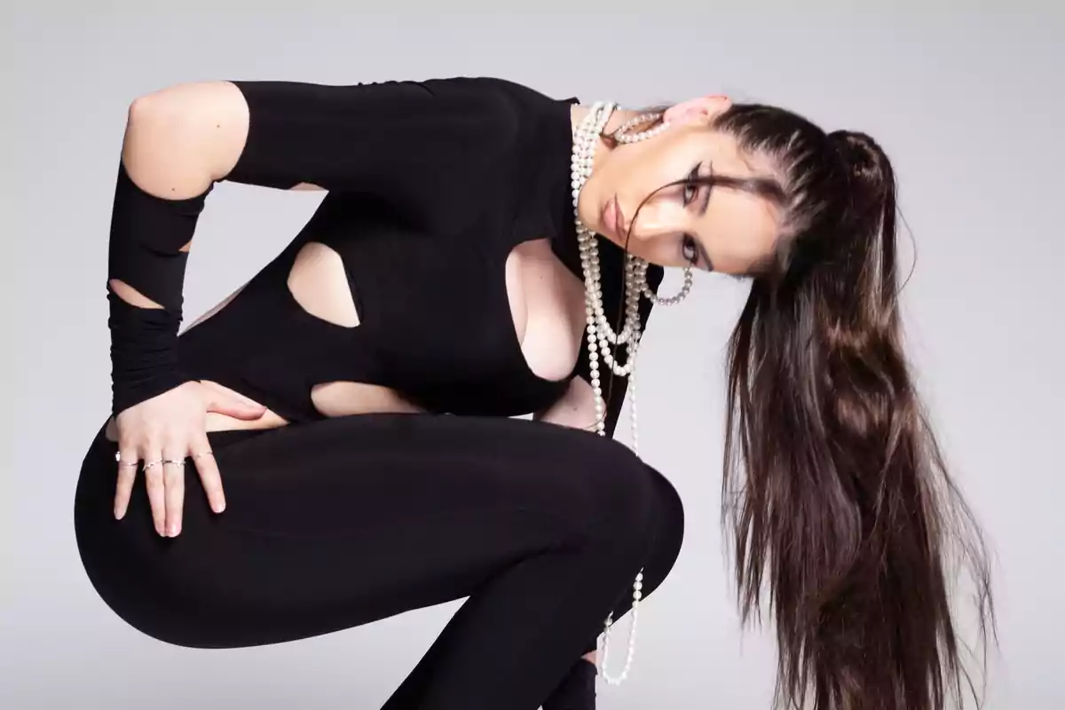 Mujer con cabello largo y oscuro vestida con un conjunto negro ajustado y accesorios de perlas posando de manera artística sobre fondo gris Mujer con cabello largo y oscuro vestida con un conjunto negro ajustado y accesorios de perlas posando de manera artística sobre fondo gris