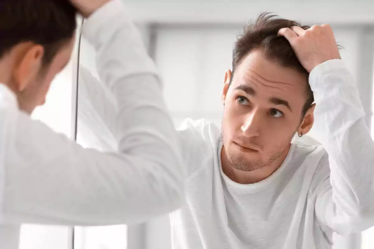 Hombre joven con camiseta blanca mirándose al espejo mientras se toca el cabello con una expresión preocupada Hombre joven con camiseta blanca mirándose al espejo mientras se toca el cabello con una expresión preocupada