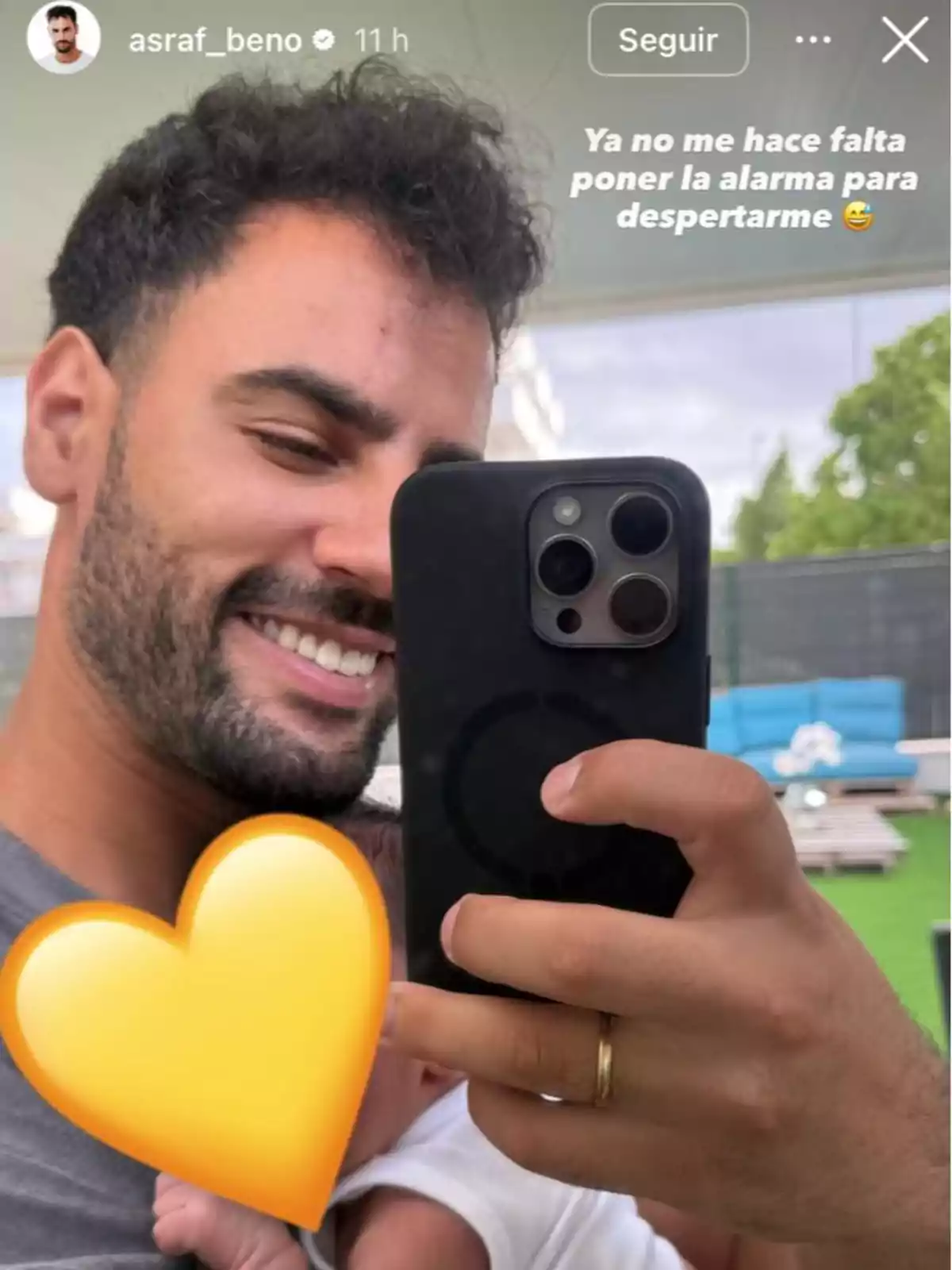 Asraf Beno sonriente tomándose una selfie con un bebé en brazos cubierto parcialmente por un emoji de corazón amarillo Asraf Beno sonriente tomándose una selfie con un bebé en brazos cubierto parcialmente por un emoji de corazón amarillo