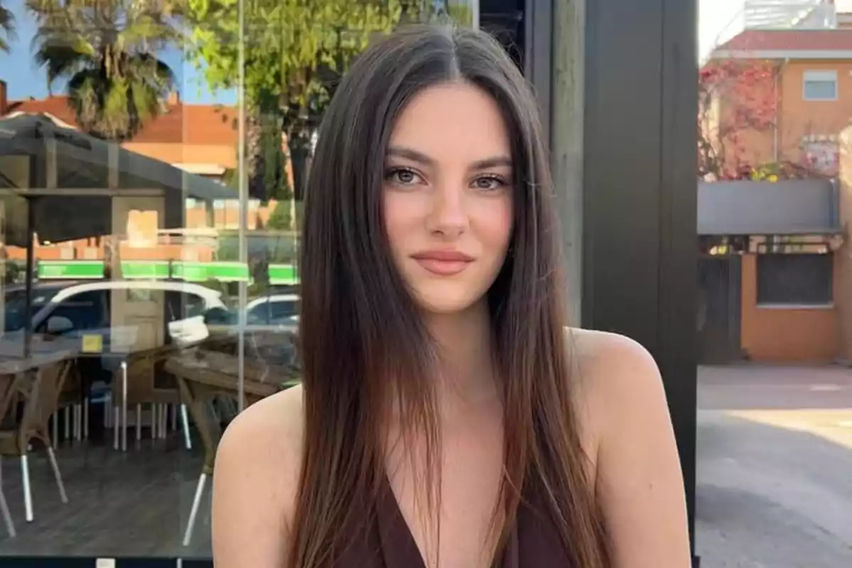 Joven de cabello largo y lacio posando al aire libre frente a una cafetería con ventanales y reflejo de autos y árboles