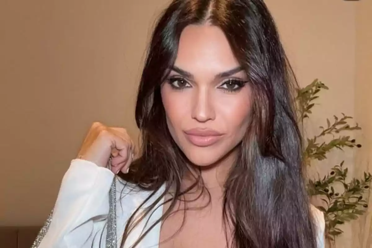Mujer de cabello largo y oscuro con maquillaje elegante, vistiendo ropa blanca y posando frente a una pared beige con una planta al fondo