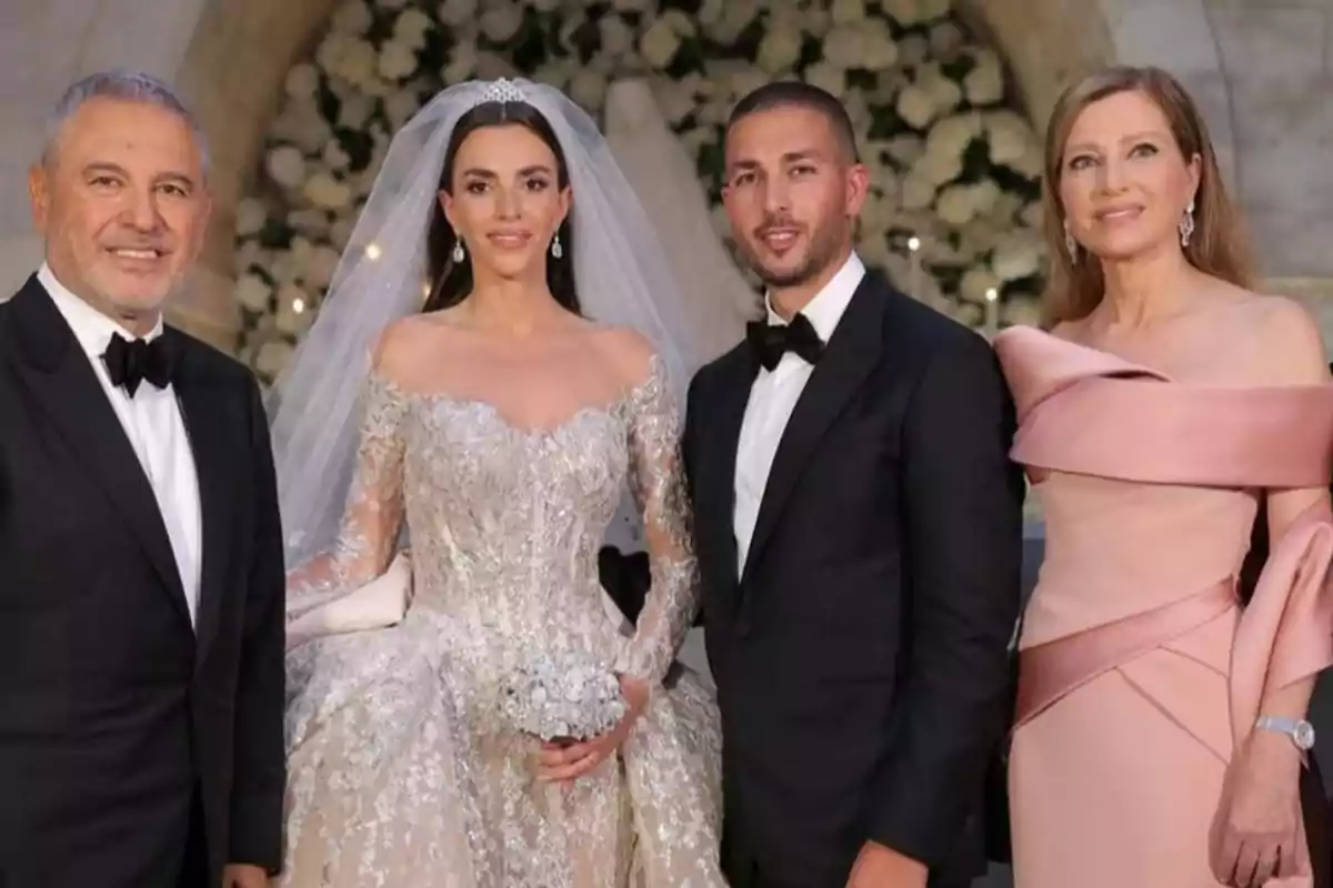 Cuatro personas elegantemente vestidas posan juntas en una boda, la novia lleva un vestido blanco con encaje y velo, el novio y otro hombre usan esmoquin negro, y una mujer lleva un vestido largo color rosa claro, al fondo se ven flores blancas.