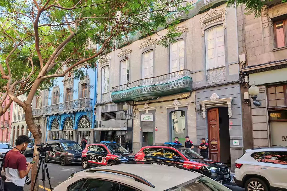 Fachada de edificios antiguos en una calle con árboles, varios autos de policía estacionados y agentes conversando en la acera mientras un camarógrafo graba la escena. Fachada de edificios antiguos en una calle con árboles, varios autos de policía estacionados y agentes conversando en la acera mientras un camarógrafo graba la escena.