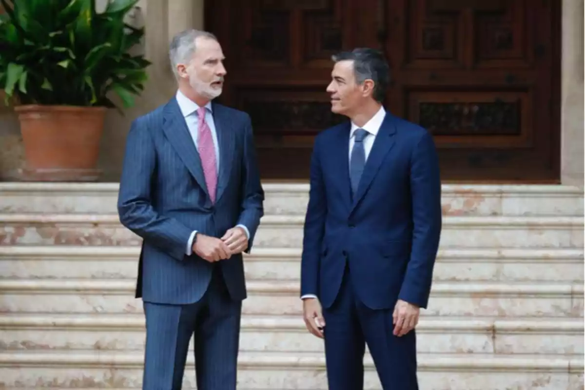 Dos hombres con traje conversan de pie frente a unas escaleras de mármol y una puerta de madera Dos hombres con traje conversan de pie frente a unas escaleras de mármol y una puerta de madera