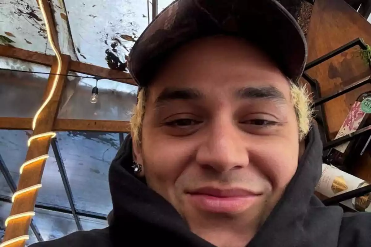 Persona joven con cabello rubio y gorra oscura sonriendo levemente en un lugar techado con decoración de luces y botellas al fondo