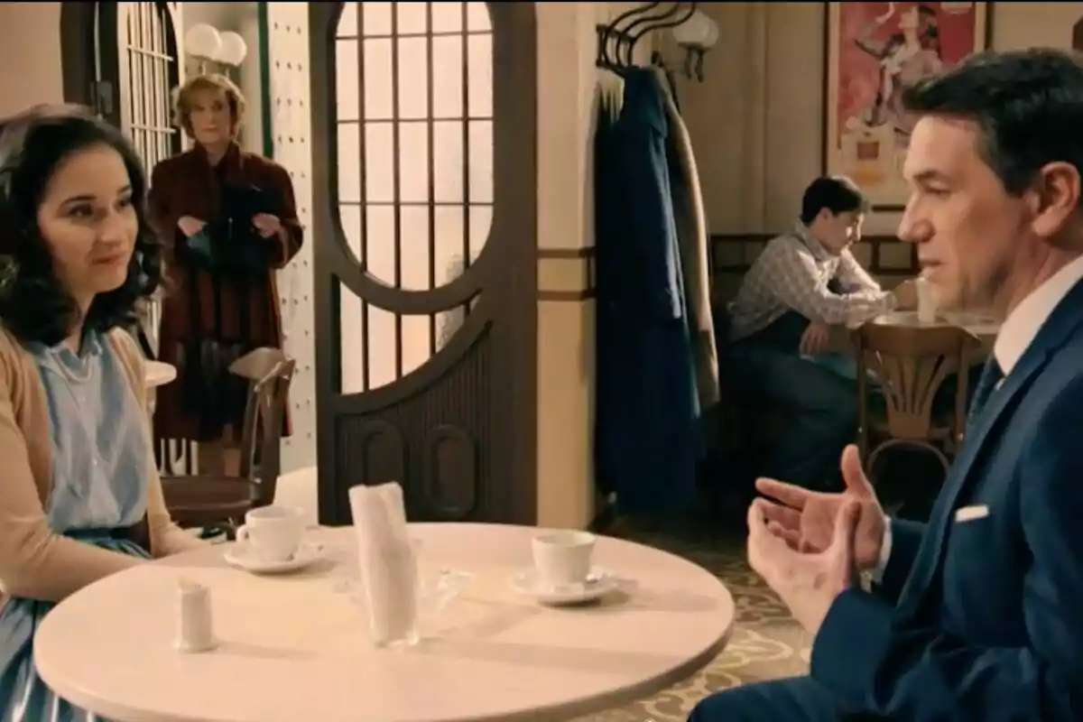 Dos personas, que son Gabriel y Cristina, conversan en una mesa de cafetería mientras una mujer observa desde el fondo y un hombre está sentado solo en otra mesa. Dos personas, que son Gabriel y Cristina, conversan en una mesa de cafetería mientras una mujer observa desde el fondo y un hombre está sentado solo en otra mesa.