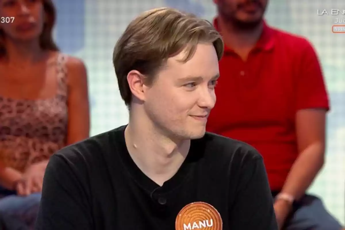 Manu de cabello claro y camiseta negra con una etiqueta que dice Manu participa en un programa de televisión mientras otras personas están sentadas al fondo. Manu de cabello claro y camiseta negra con una etiqueta que dice Manu participa en un programa de televisión mientras otras personas están sentadas al fondo.