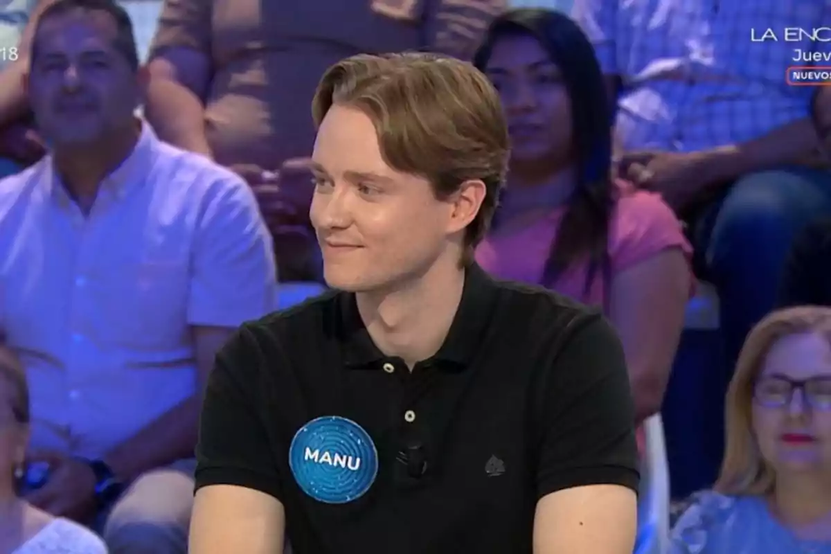 Manu con camiseta negra y una placa azul que dice Manu participa en un programa de concursos con público al fondo. Manu con camiseta negra y una placa azul que dice Manu participa en un programa de concursos con público al fondo.