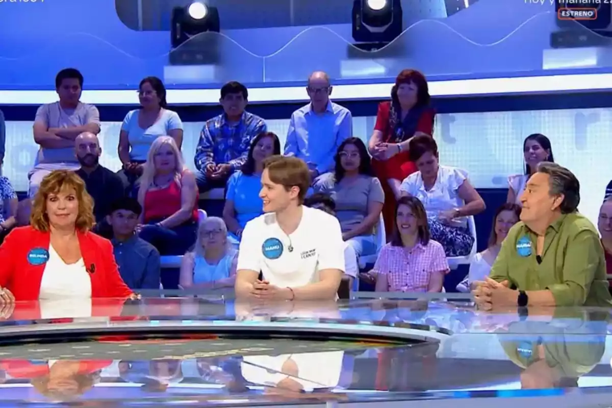 Tres personas sentadas en una mesa de un programa de concursos con público al fondo Tres personas sentadas en una mesa de un programa de concursos con público al fondo