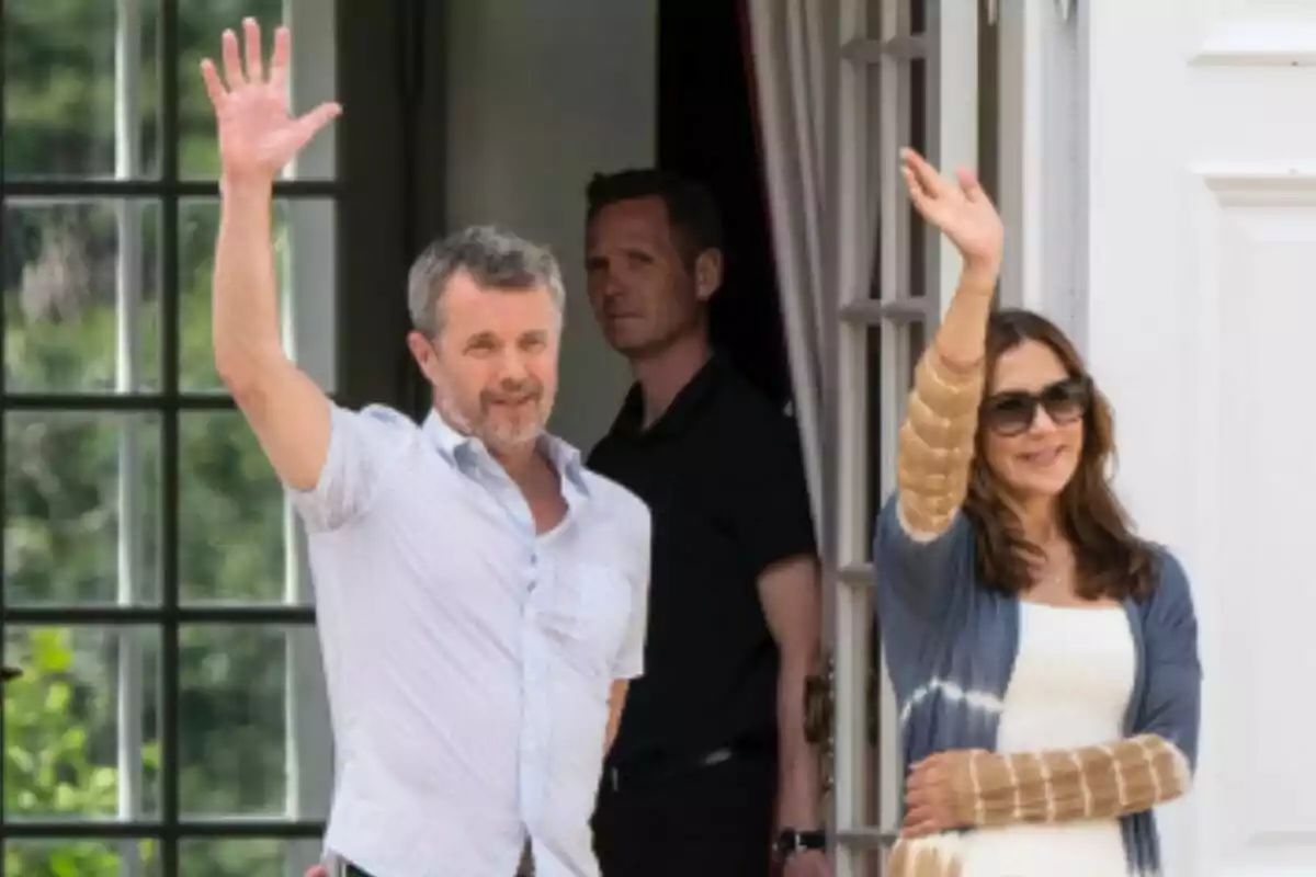 Una pareja sonriente -formada por Mary de Dinamarca y Federico de Dinamarca- saluda con la mano levantada desde la entrada de un edificio mientras una persona de fondo los observa. Una pareja sonriente -formada por Mary de Dinamarca y Federico de Dinamarca- saluda con la mano levantada desde la entrada de un edificio mientras una persona de fondo los observa.