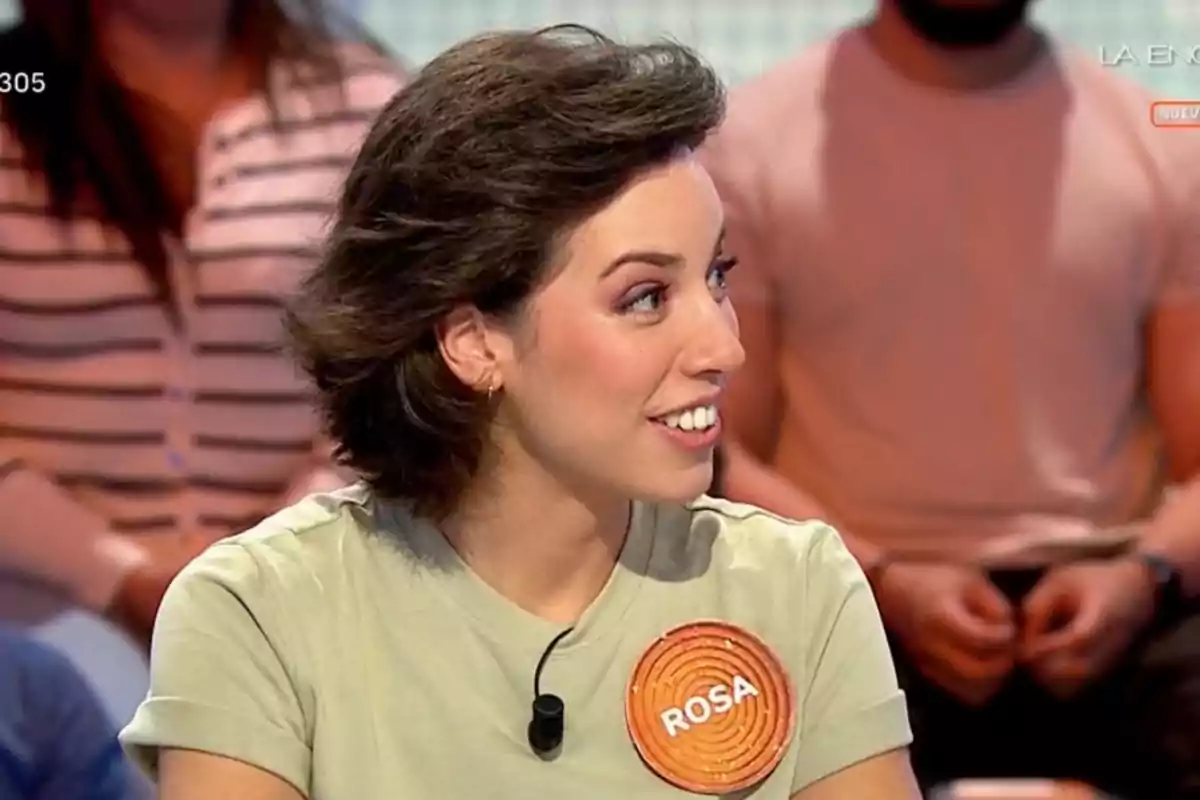 Rosa joven con cabello corto y oscuro, lleva una camiseta clara y una chapa naranja con el nombre Rosa, está sentada en un plató de televisión con otras personas al fondo. Rosa joven con cabello corto y oscuro, lleva una camiseta clara y una chapa naranja con el nombre Rosa, está sentada en un plató de televisión con otras personas al fondo.