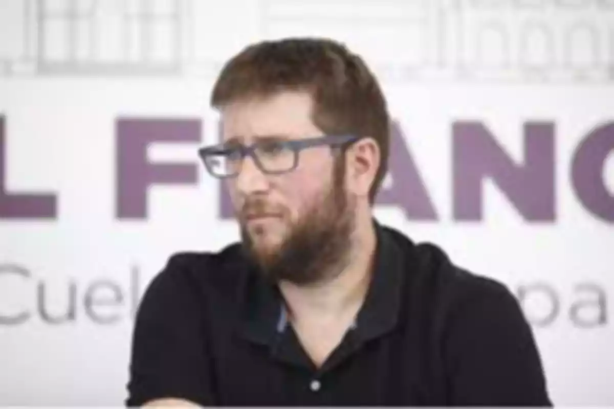 Hombre con barba y gafas sentado frente a un fondo borroso con letras grandes Hombre con barba y gafas sentado frente a un fondo borroso con letras grandes