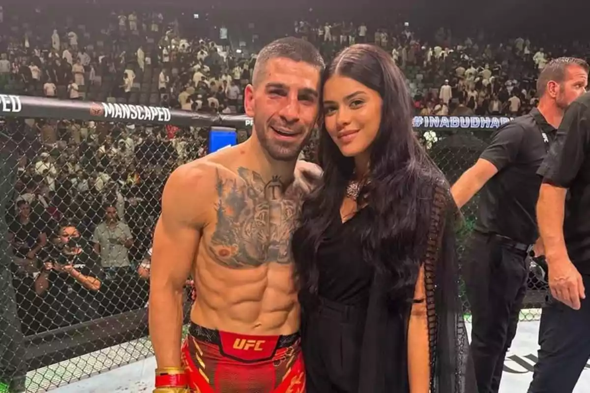 Un peleador de artes marciales mixtas sin camiseta y con pantalones cortos de la UFC posa sonriente junto a una mujer de cabello largo y oscuro dentro de un octágono, con público al fondo. Un peleador de artes marciales mixtas sin camiseta y con pantalones cortos de la UFC posa sonriente junto a una mujer de cabello largo y oscuro dentro de un octágono, con público al fondo.
