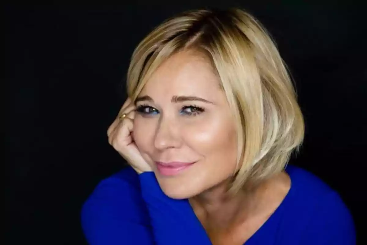 Mujer rubia de cabello corto con suéter azul sonriendo y apoyando la cabeza en su mano frente a un fondo negro