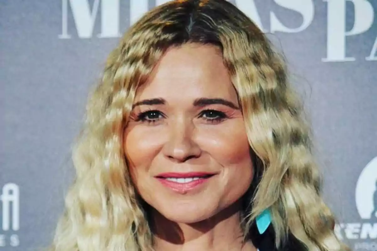 Mujer de cabello rubio ondulado sonriendo frente a un fondo gris en un evento