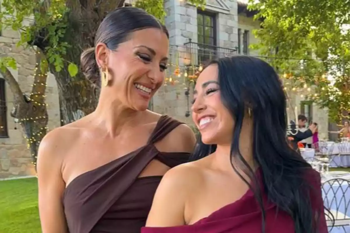 Dos mujeres sonríen y se miran con cariño en un evento al aire libre rodeadas de árboles y luces decorativas